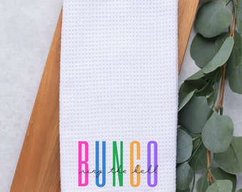 Peut inclure: Serviette de cuisine en tissu gaufré blanc avec le texte "BUNCO" en couleurs arc-en-ciel et le texte "ring the bell" en écriture cursive noire.