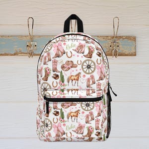 May include: White backpack with a repeating pattern of cowboy boots, horses, hats, horseshoes, and pink bows. The word "Rubie" is printed on the backpack. Features a black handle, black zipper, and a mesh side pocket.