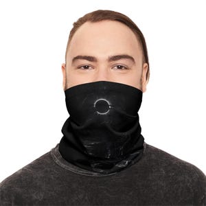 Può includere: Scaldacollo nero con un design circolare bianco su uno sfondo scuro e strutturato. Lo scaldacollo è indossato attorno al collo e copre la parte inferiore del viso. Il design ha un tema celeste o cosmico.