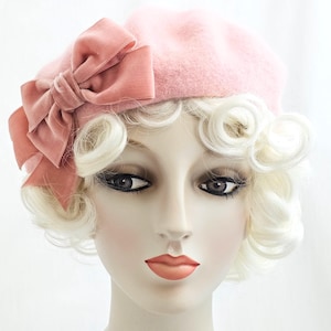 Op de afbeelding: Een lichtroze baret met een grote, bijpassende roze fluwelen strik aan de zijkant. De hoed is gemaakt van zacht viltmateriaal en rust op een etalagepophoofd met kort, krullend blond haar en geschilderde gelaatstrekken.
