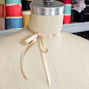 May include: A beige satin ribbon tied in a bow around the neck of a dress form. The dress form is a light beige color. The background features spools of colorful ribbon.