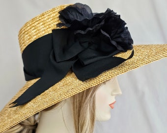 Puede incluir: Un sombrero de paja de ala ancha con una cinta negra y una gran flor de tela negra. El sombrero es de color paja natural y tiene una ala ancha, adecuado para la protección solar. La cinta negra rodea la corona.