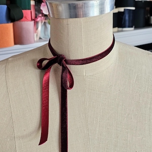 Puede incluir: Un collar gargantilla de terciopelo burdeos con un lazo atado en la parte delantera. El collar está en un maniquí de tela blanca.