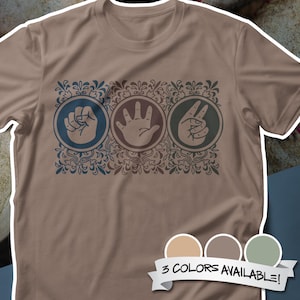 May include: A tan t-shirt featuring a graphic of the rock, paper, scissors hand gestures in blue, brown, and green circles. The design is framed by a decorative floral border. The shirt has a crew neck and short sleeves.