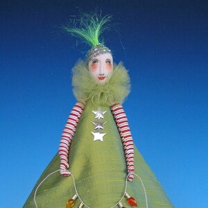 Pode incluir: Uma boneca de árvore de Natal verde com uma estrela prateada, um colar de tule verde com babados e uma corda de luzes coloridas. A boneca tem um rosto branco com traços pintados e cabelo verde. A boneca está em pé sobre uma base de madeira.