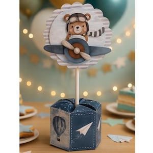 Pode incluir: Decoração de festa azul e branca com um urso com chapéu e cachecol de piloto, sentado em um avião com hélice. A decoração está em um palito, colocada em uma caixa azul com um design de balão de ar quente e avião de papel. A palavra "PARTOON" está na parte inferior.