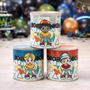 Puede incluir: Tres recipientes metálicos coloridos con tapas, cada uno con un personaje de dibujos animados sosteniendo un mando de videojuegos. Los recipientes están decorados con gráficos y texto temáticos de juegos, con una tarta de cumpleaños en el fondo.