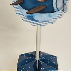 May include: A whimsical blue and brown airplane-themed centerpiece. The design features a cartoon bear pilot in a blue plane, set against a cloud and striped background. The base is a blue box with paper airplane and heart details.