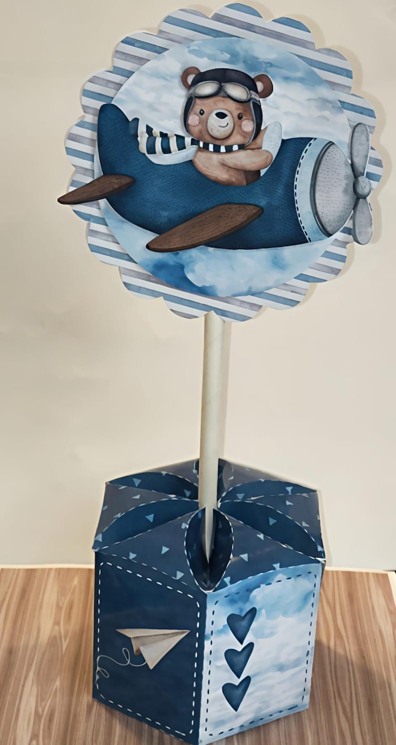 May include: A whimsical blue and brown airplane-themed centerpiece. The design features a cartoon bear pilot in a blue plane, set against a cloud and striped background. The base is a blue box with paper airplane and heart details.