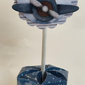 May include: A whimsical centerpiece featuring a cartoon bear in a pilot's hat and scarf, piloting a blue and brown airplane. The design is set against a striped background and sits atop a blue box with a hot air balloon and paper airplane illustration.