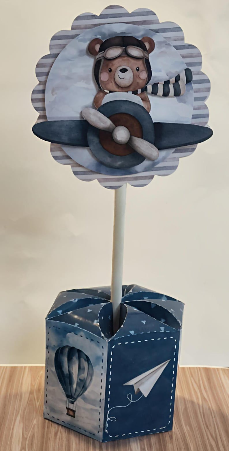 May include: A whimsical centerpiece featuring a cartoon bear in a pilot's hat and scarf, piloting a blue and brown airplane. The design is set against a striped background and sits atop a blue box with a hot air balloon and paper airplane illustration.