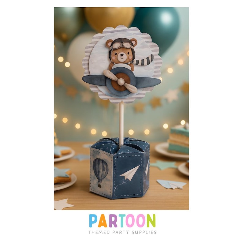 May include: A blue and white party decoration featuring a bear in a pilot's hat and scarf, sitting in a plane with a propeller. The decoration is on a stick, placed in a blue box with a hot air balloon and paper airplane design. The word "PARTOON" is at the bottom.