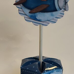 May include: A decorative centerpiece featuring a blue airplane with a teddy bear pilot. The airplane is mounted on a white stick, which is inserted into a blue box with a paper airplane design and heart accents. The design is suitable for a baby shower or birthday.
