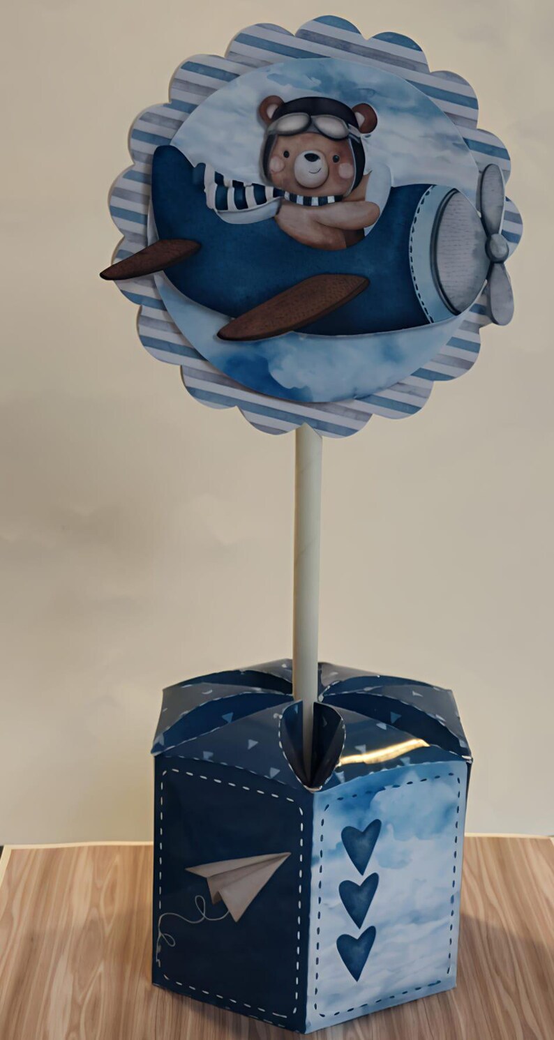 May include: A decorative centerpiece featuring a blue airplane with a teddy bear pilot. The airplane is mounted on a white stick, which is inserted into a blue box with a paper airplane design and heart accents. The design is suitable for a baby shower or birthday.