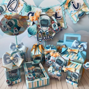 May include: A blue and yellow astronaut-themed party decoration set with a banner, boxes, and a centerpiece. The banner says "Davi" and features stars, planets, and a rocket. The boxes are decorated with astronauts, stars, and planets. The centerpiece is a round cake topper with an astronaut and a rocket.