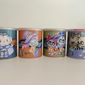 Op de afbeelding: Vier decoratieve metalen containers met Halloween-thema illustraties. De ontwerpen omvatten een vampier, een heks op een bezem, een stel in Day of the Dead-kleding en een skelet met een wolf. Elke container heeft een ander kleurenschema.
