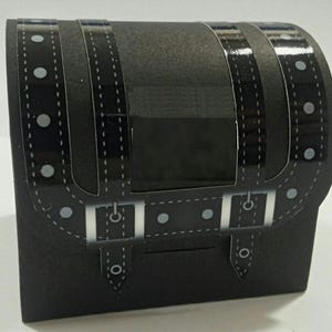 May include: A black cardboard box shaped like a treasure chest with a faux leather design and silver buckles. The box has a flap that opens to reveal the inside.