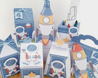 May include: A set of party favor boxes decorated with an astronaut and rocket ship theme. The boxes are blue, white, and orange with a grid pattern. The boxes have the name "Matheus" on them.