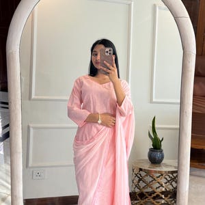 May include: A light pink, three-piece outfit, reflected in a mirror. The outfit includes a long-sleeved top, pants, and a draped shawl. The outfit is made of a soft, flowing fabric. The person is wearing silver sandals and a gold watch.