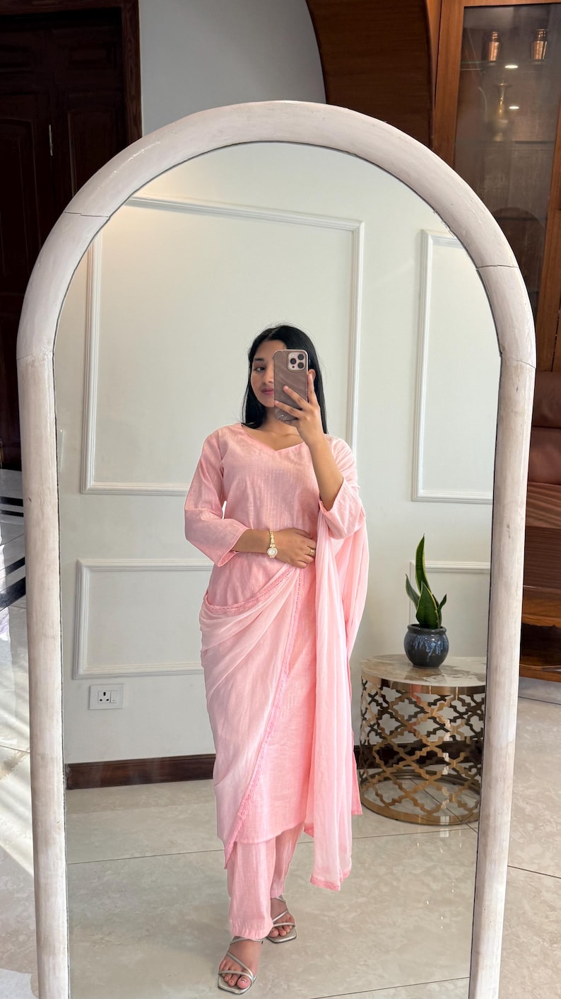 May include: A light pink, three-piece outfit, reflected in a mirror. The outfit includes a long-sleeved top, pants, and a draped shawl. The outfit is made of a soft, flowing fabric. The person is wearing silver sandals and a gold watch.