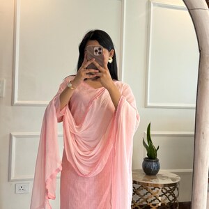 May include: A light pink salwar kameez suit with a matching dupatta. The suit features a straight-cut kurta and pants, with a dupatta draped over the shoulders. The edges of the dupatta and kurta are trimmed with a darker pink border.