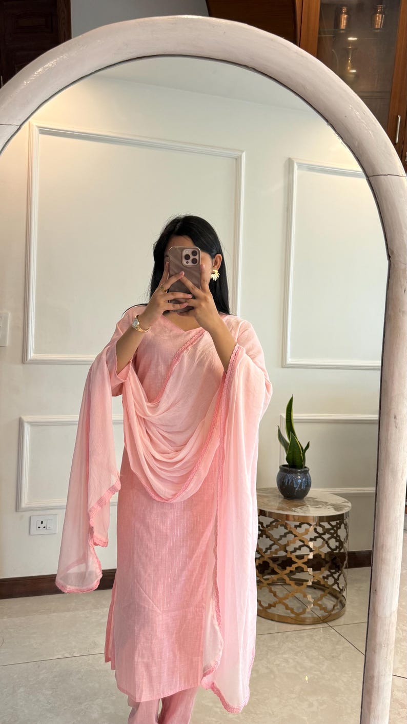 May include: A light pink salwar kameez suit with a matching dupatta. The suit features a straight-cut kurta and pants, with a dupatta draped over the shoulders. The edges of the dupatta and kurta are trimmed with a darker pink border.
