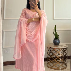 May include: A light pink salwar kameez set, including a tunic, pants, and a draped scarf. The outfit features delicate embroidery along the edges and is made of a lightweight fabric. A small side table with a marble top and a decorative plant is visible.