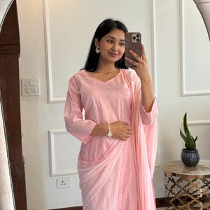 May include: A light pink, three-piece outfit consisting of a long-sleeved top, draped skirt, and matching pants. The top has a V-neck and the skirt is draped over the pants. The outfit is made of a lightweight fabric. The outfit is worn with a gold watch and earrings.