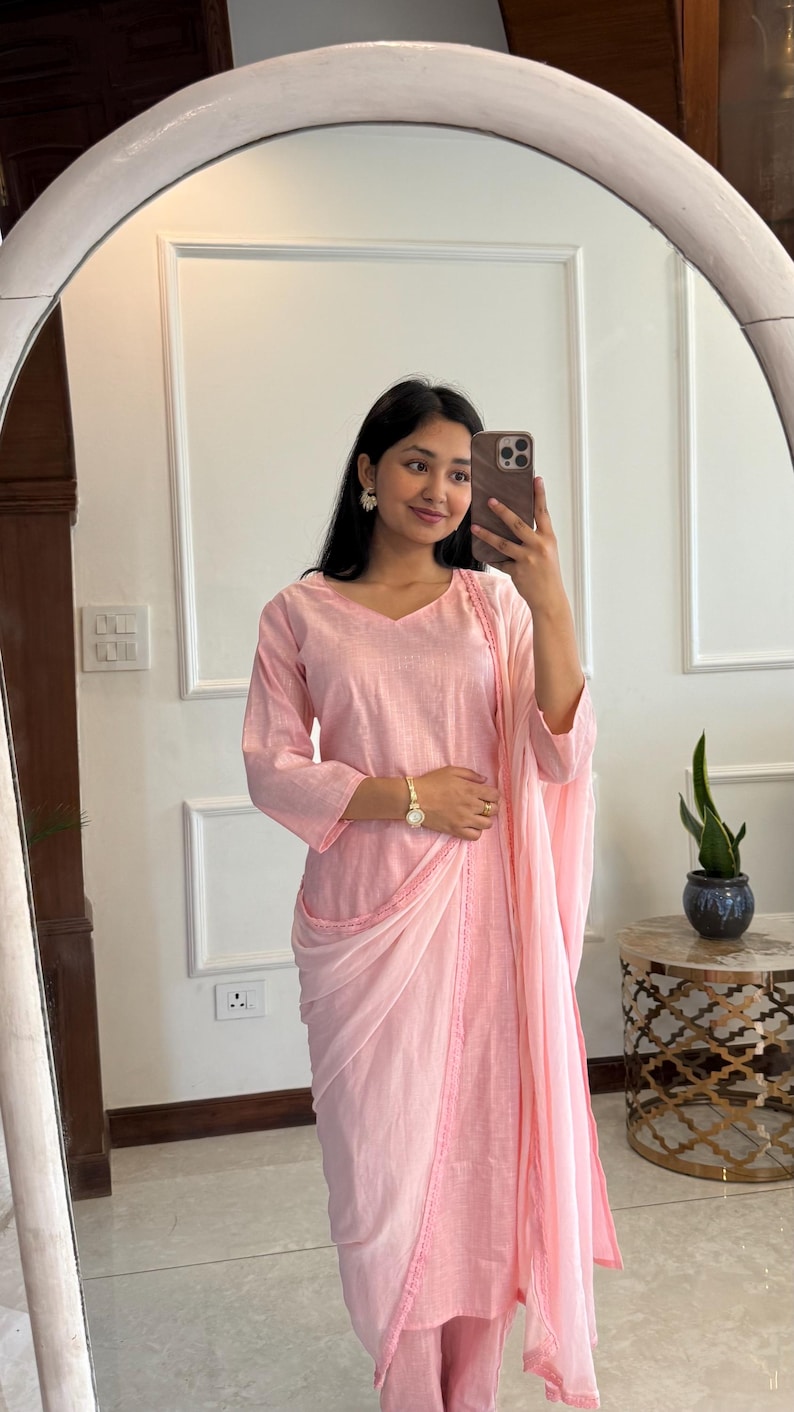 May include: A light pink, three-piece outfit consisting of a long-sleeved top, draped skirt, and matching pants. The top has a V-neck and the skirt is draped over the pants. The outfit is made of a lightweight fabric. The outfit is worn with a gold watch and earrings.