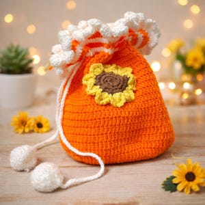 Op de afbeelding: Een oranje gehaakte trekkoordtas met een zonnebloem applicatie. De tas heeft een wit en oranje gerimpelde rand en een wit trekkoord met twee witte pompons. De tas staat op een houten oppervlak.