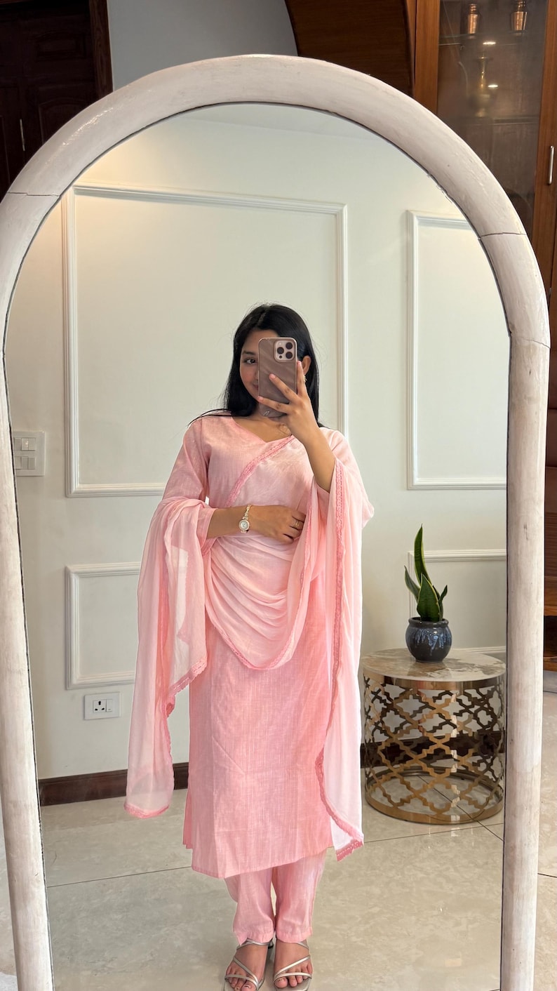 May include: A light pink salwar kameez set, including a tunic, pants, and a dupatta. The tunic and dupatta have delicate pink trim. The outfit is made of a lightweight fabric. The person in the image is wearing silver sandals.