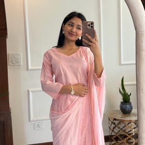 May include: A light pink, long-sleeved outfit with a draped shawl. The outfit includes a V-neck top and matching pants. The outfit is made of a soft fabric and is suitable for formal or casual wear. The outfit is reflected in a large, white-framed mirror.