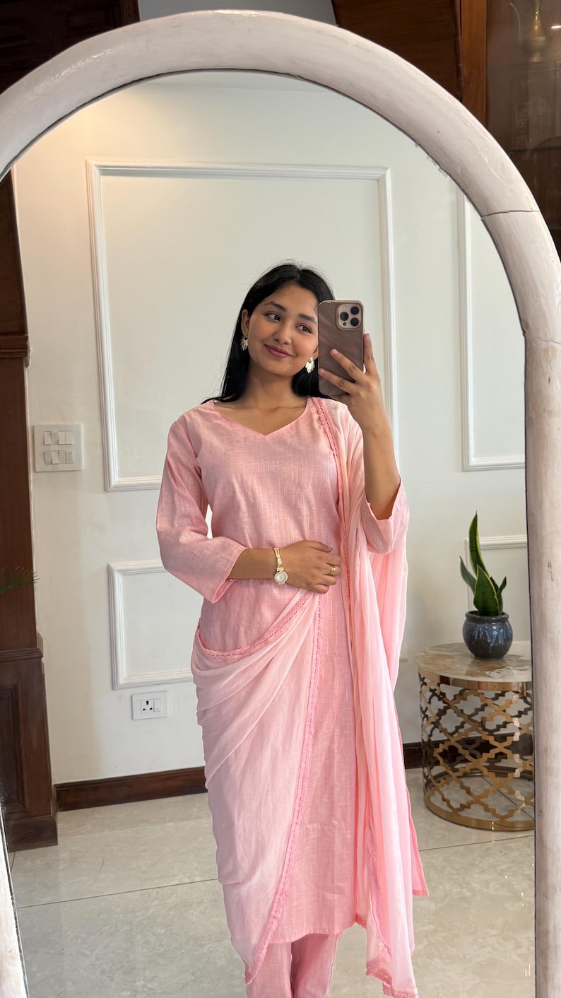 May include: A light pink, long-sleeved outfit with a draped shawl. The outfit includes a V-neck top and matching pants. The outfit is made of a soft fabric and is suitable for formal or casual wear. The outfit is reflected in a large, white-framed mirror.