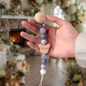 May include: A wooden baby pacifier clip with the name "Noah" engraved on a bear-shaped charm. The clip features a wooden bead, geometric and round beads in shades of gray, and a white bead at the end.