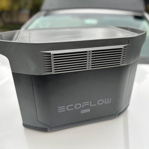 May include: A dark gray portable power station with the brand name ECOFLOW MAX printed on the front. The unit has a rectangular shape with a flat top and a vent with horizontal slats. It is sitting on a white surface.