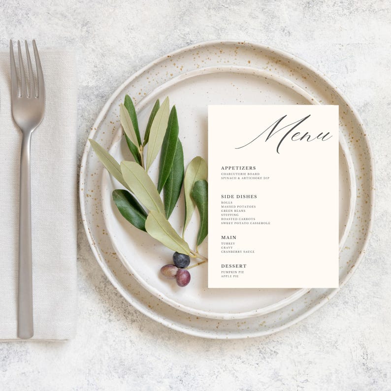 May include: A simple, minimalist menu card with a handwritten font. The menu lists appetizers, side dishes, main course, and dessert. The card is on a white plate with a green olive branch and dark blue berries.