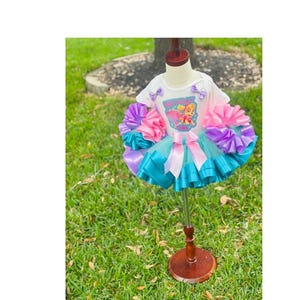 May include: A white t-shirt with a butterfly and a character from the cartoon show 'Super Wings' on it. The shirt is paired with a turquoise tulle tutu with pink and purple ribbon accents.