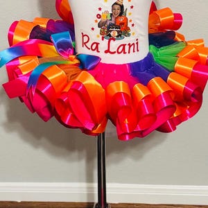 May include: A colorful birthday outfit for a child, featuring a white t-shirt with a graphic and the name "Ra Lani" in red. The outfit includes a rainbow tutu skirt and matching bow.