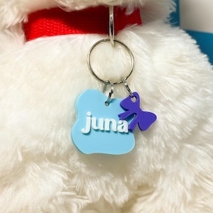 May include: A light blue, cloud-shaped pet ID tag featuring the white text juna, attached to a red collar with a silver metal ring. A small, purple, bow-shaped charm hangs from the same ring, resting against the white, plush, furry texture.