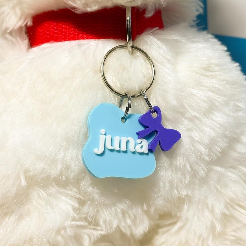 May include: A light blue, cloud-shaped pet ID tag featuring the white text juna, attached to a red collar with a silver metal ring. A small, purple, bow-shaped charm hangs from the same ring, resting against the white, plush, furry texture.