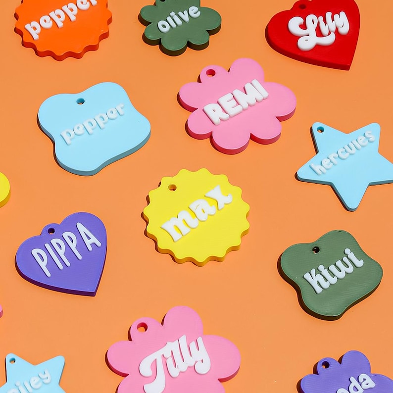 May include: A collection of colorful acrylic pet tags in various shapes including hearts, stars, flowers, and scalloped circles. Each tag features raised white lettering with names such as pepper, olive, Lily, REMI, hercules, max, PIPPA, Kiwi, and Tilly.