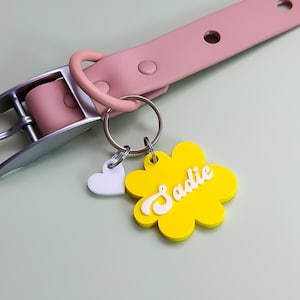 May include: A light pink collar with a silver metal buckle and a metal ring attached to a yellow flower-shaped tag. The tag features the name Sadie in white script lettering and includes a small white heart-shaped charm hanging from the ring.