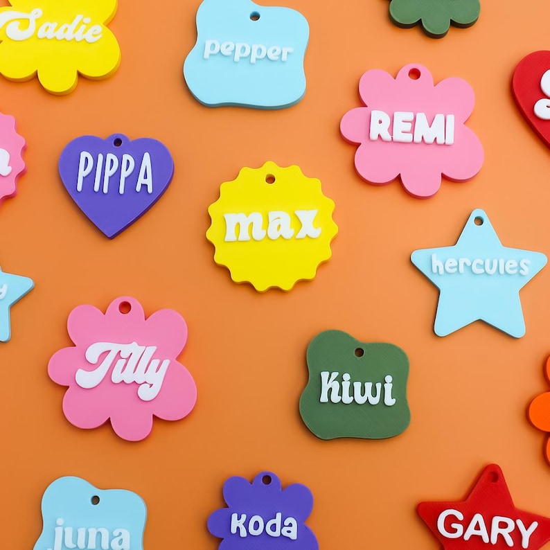 May include: A collection of colorful acrylic pet tags in various shapes including flowers, hearts, and stars. Each tag features a unique name in raised white lettering, such as Sadie, Pepper, Remi, Pippa, Max, Hercules, Tilly, Kiwi, Juna, Koda, and Gary.