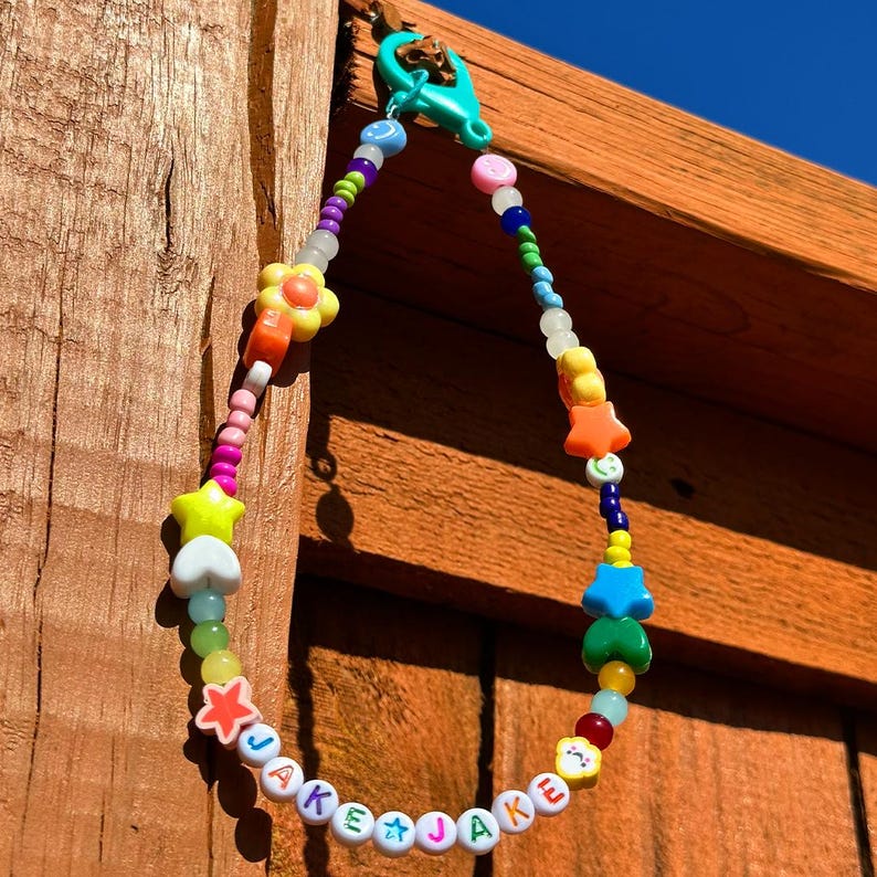 May include: A colorful beaded keychain with the name "JAKE" spelled out in white letters. The keychain features a variety of colorful beads, including stars, hearts, flowers, and smiley faces.
