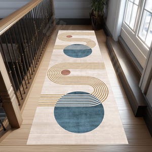 May include: A rectangular beige runner rug with a geometric design. The rug features curved lines in tan and brown, along with teal and brown circles. The rug is placed on a wooden floor near a staircase and a doorway.