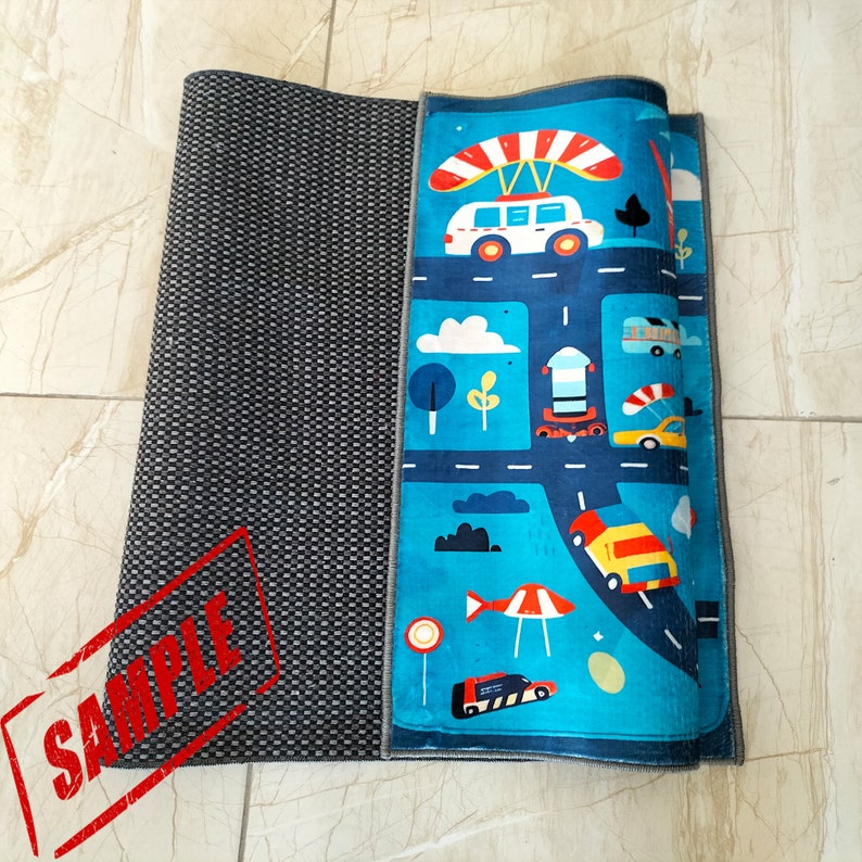 May include: A blue and gray play mat with a colorful design featuring cars, trucks, and a hot air balloon. The mat has a gray border and a black and white checkered pattern on the left side.
