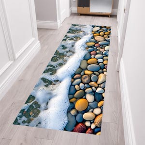 Puede incluir: Una alfombra rectangular con un diseño de escena de playa. La alfombra presenta una imagen detallada de guijarros coloridos y olas oceánicas espumosas. La paleta de colores incluye tonos de azul, marrón, amarillo y blanco. La alfombra está diseñada para parecerse a una costa.