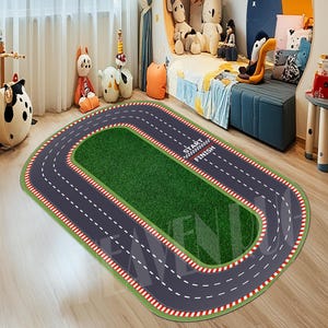 May include: A green and gray oval-shaped race track rug with white lines and red and white checkered start and finish lines. The rug has the words "Start" and "Finish" printed on it.