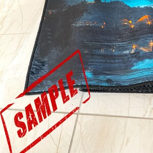 K&ouml;nnte beinhalten: Nahaufnahme eines schwarzen und blauen Teppichs mit einem Stadtbild-Design. Der Teppich hat einen Fransenrand und das Wort "SAMPLE" ist rot auf das Bild gestempelt.