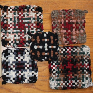 May include: Five woven pot holders in various colors and patterns. The pot holders are made of a thick, textured fabric and have a square shape. The colors include black, white, red, brown, and green.